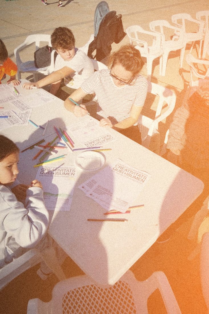 Infants i adults dibuixen a una taula amb els cartells de Creativitats que (+) sumen, a Pòrtol, Marratxí